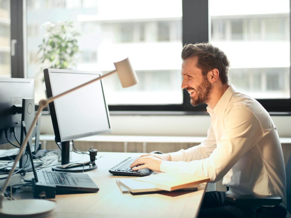 man in white dress shirt sitting on black rolling chair while facing black computer set and smiling